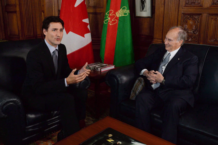 A dark-haired man gestures as he speaks in his office with an older man smiling as he looks on.