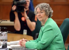 An older woman with short grey hair in a green suit jacket sits in a committee room.