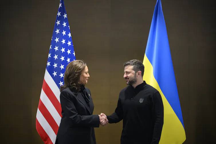 A woman and a man shake hands in front of Ukraine and US flags.