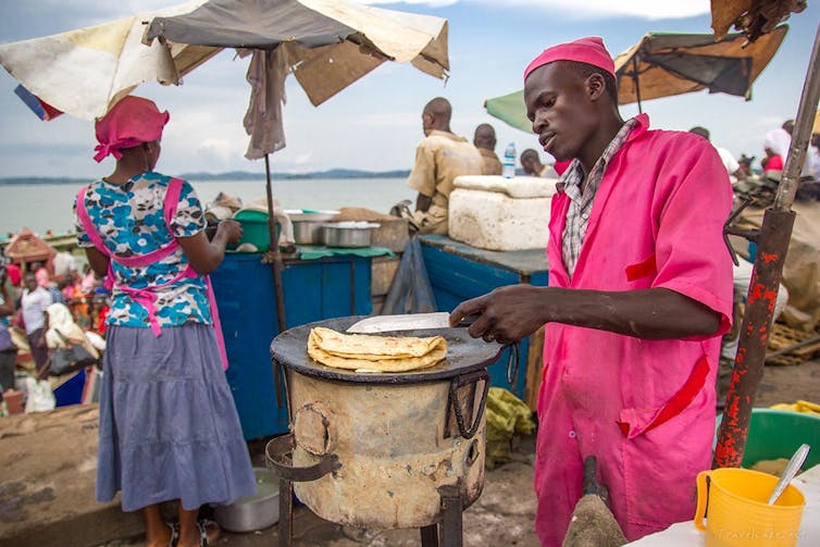 People cook in bright traditional clothing on large pans under umbrellas next to water. Aman cooks what looks like a large omelette.