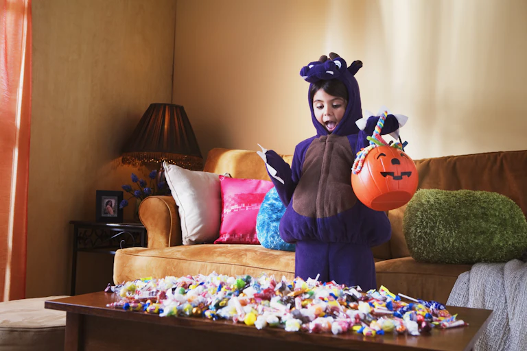 Child in costume holding pumpkin basket full of candy, gaping at a table also laden with candy