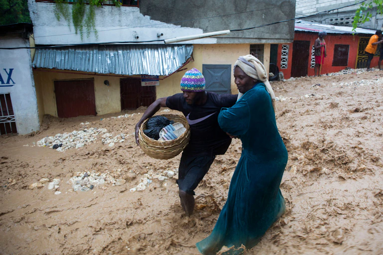 Two women struggle through a street flooding with muddy water in Haiti. One carries a basket