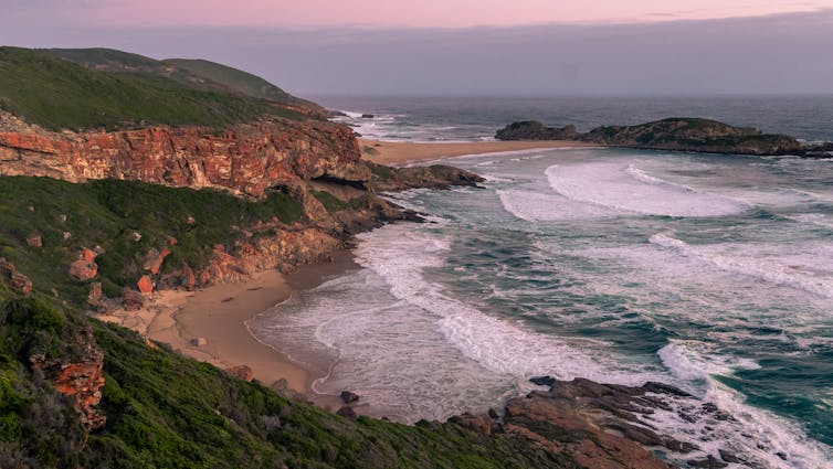 A landscape image of craggy cliffs topped with greenery against which ocean waves are lapping