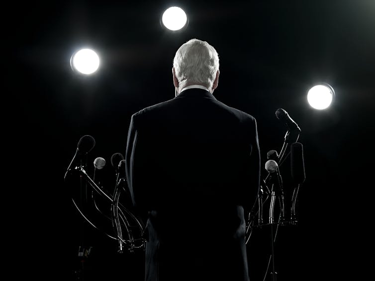 The back of a man in a suit talking into microphones with strong lights on him.