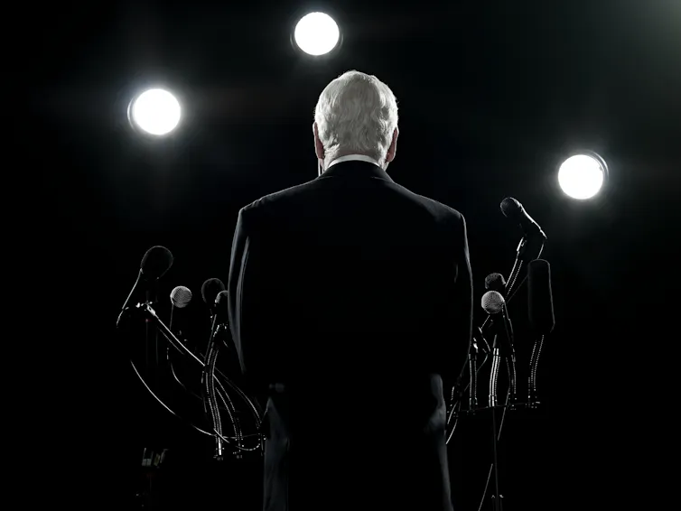 The back of a man in a suit talking into microphones with strong lights on him.