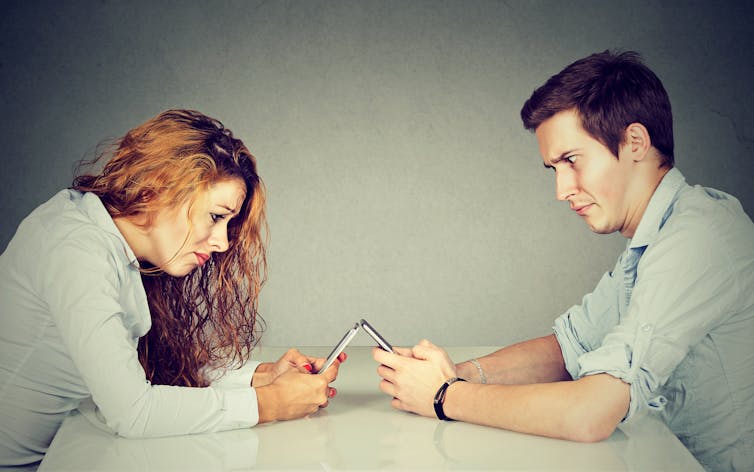A young woman and man sitting at table looking at their smartphones with frustrated expressions.