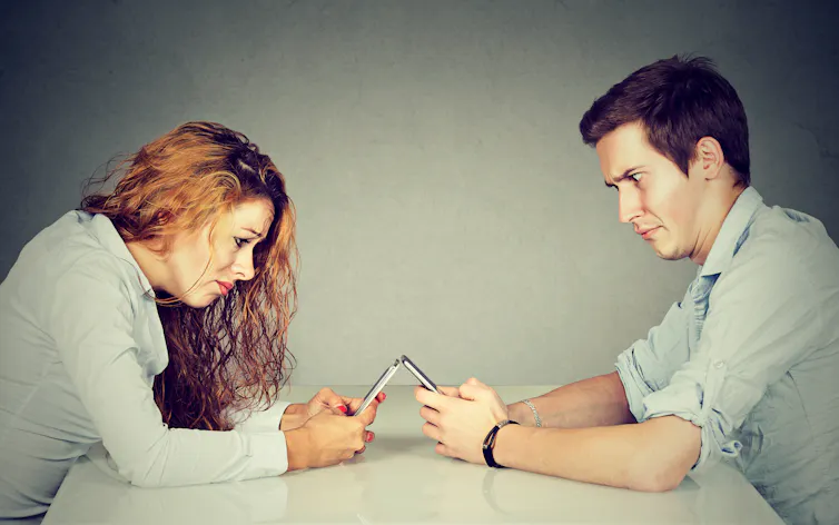 A young woman and man sitting at table looking at their smartphones with frustrated expressions.