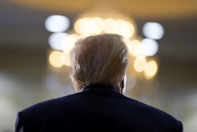 A man with fluffy grey-blond hair is photographed from behind from the shoulders up.