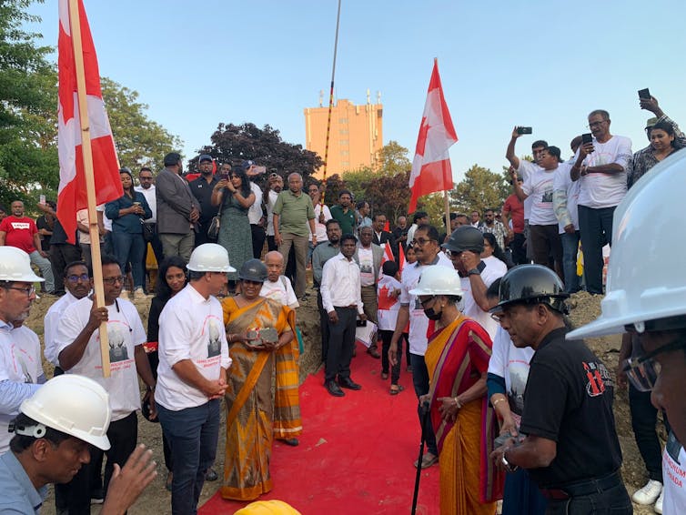 A group of people wearing hart hats stand around a red carpet. Some carry Canadian flags.
