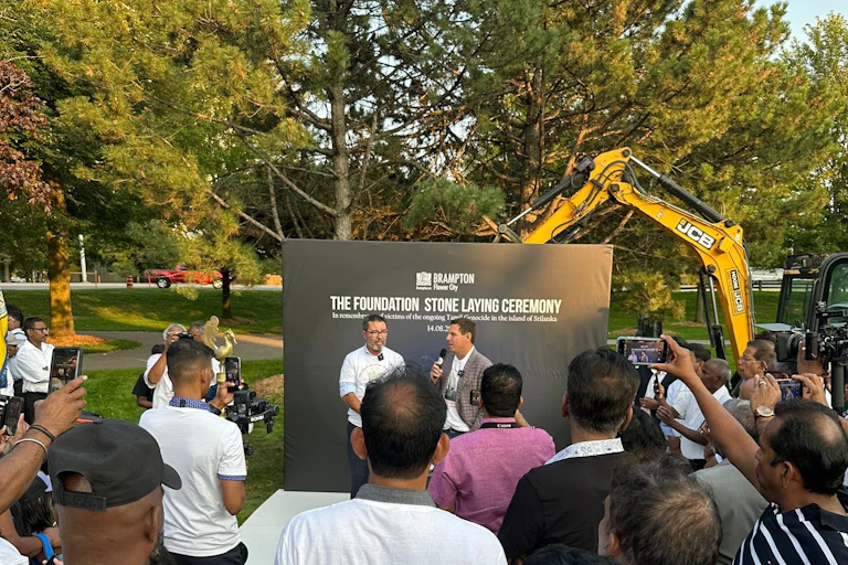 A man speaking into a mic in front of a sign reading foundation stone laying ceremony. Others are gathered around. They are standing in a park