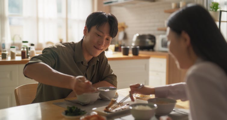 A man and woman in a light-filled kitchen use chopsticks to share from the same dish.