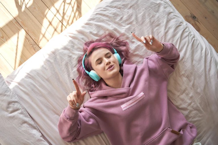A young girl with pink hair and headphones lies on a bed listening to music. She points her figures in the air.