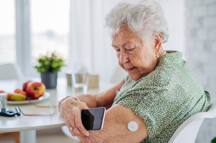 An elderly woman with a continuous glucose monitor patch looks at her phone.