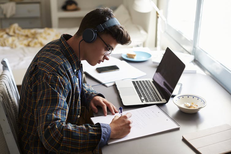 A teenage boy writes at a desk in a bedroom, he has a laptop next to him.