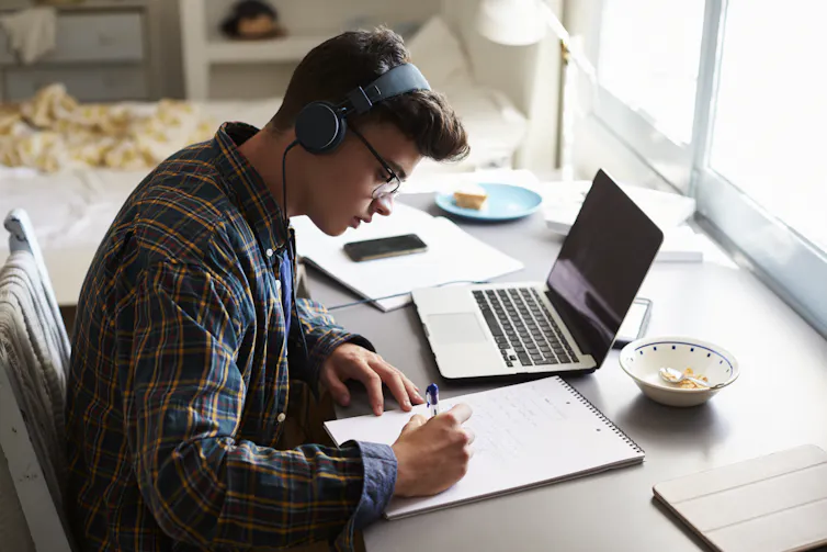 A teenage boy writes at a desk in a bedroom, he has a laptop next to him.