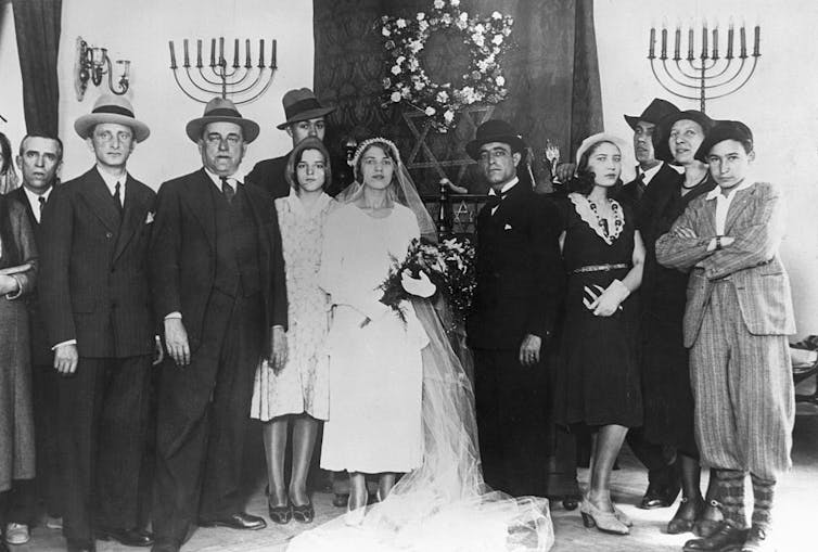 A black and white portrait of around a dozen people standing around a bride and groom, with candelabras in the background.