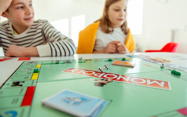 Two children sit next to a Monopoly board.