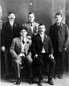 Two rows of men, the first row seated, posing for a black and white portrait while wearing suits.