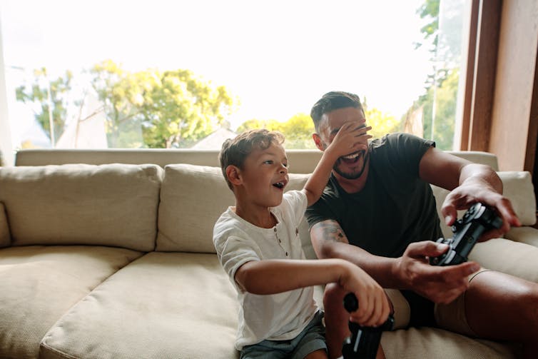 A young boy puts his hand over a man's eyes as they play a video game on the couch.