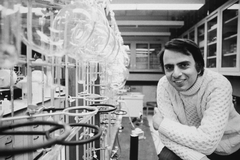 A black and white photograph of Carl Sagan in his science laboratory.