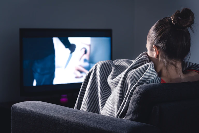 Woman on sofa, wrapped in blanket watching TV horror movie