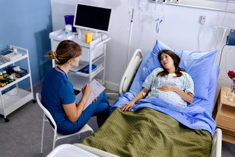 Female nurse taking notes and talking to a pregnant woman in a hospital bed.