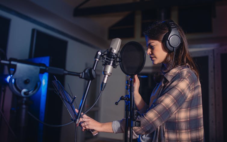 A young white woman in a professional recording studio with headphones on standing at a mic.