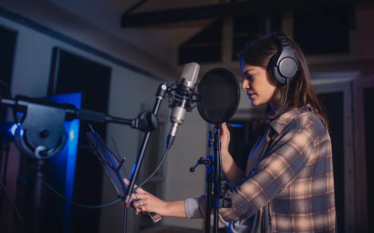 A young white woman in a professional recording studio with headphones on standing at a mic.