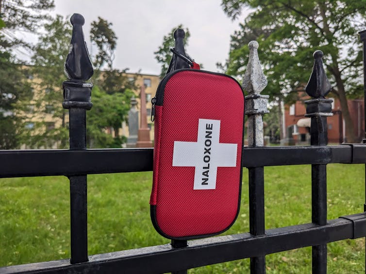 A pouch containing the drug Naloxone hangs on an iron fence.
