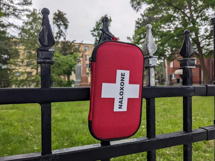 A pouch containing the drug Naloxone hangs on an iron fence.