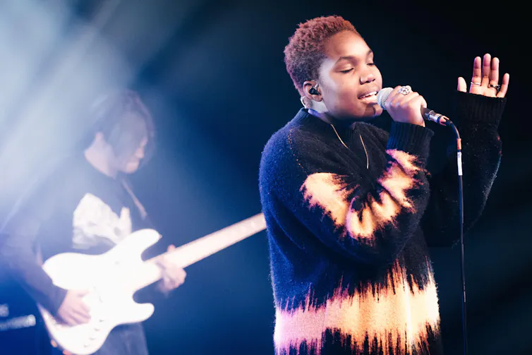 A young black female singer on stage with a guitarist standing behind.