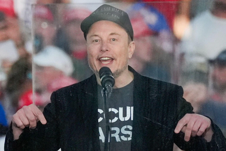 Elon Musk stands in front of a monitor speaking at a Trump rally.