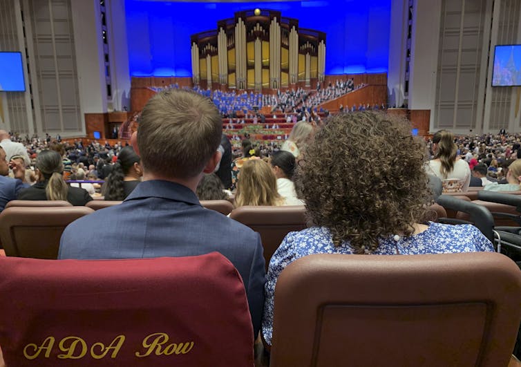 A man and a woman, seated with their backs to the camera, look toward a large organ on stage in an auditorium.