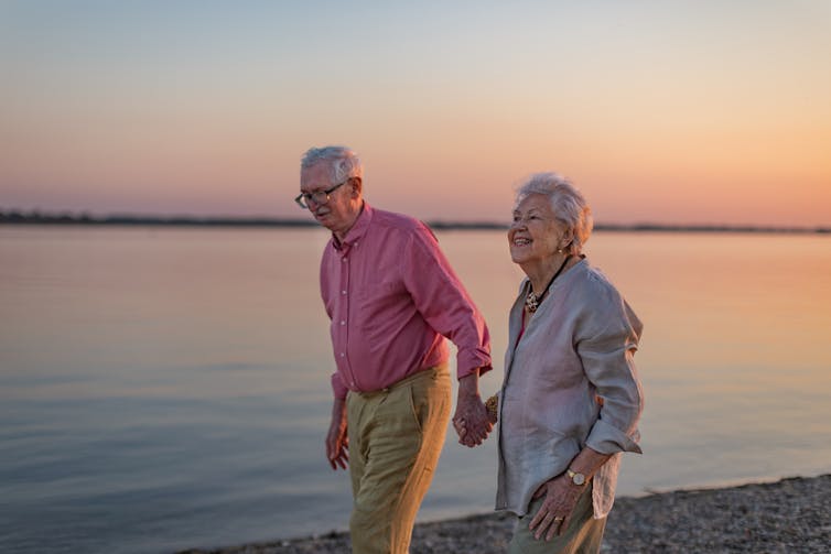 A couple, holding hands, looking happy, stroll by the sea as the sun sets.
