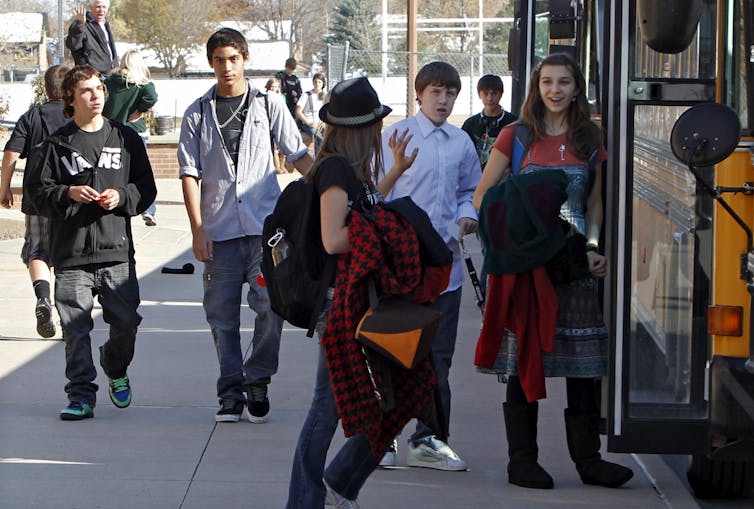 A group of middle schoolers greet each other on the sidewalk next to a yellow school bus.