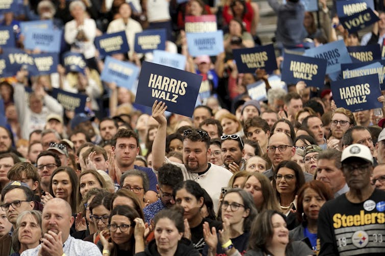 People hold Harris Walz signs. One man wears. Pittsburgh Steelers T-shirt.