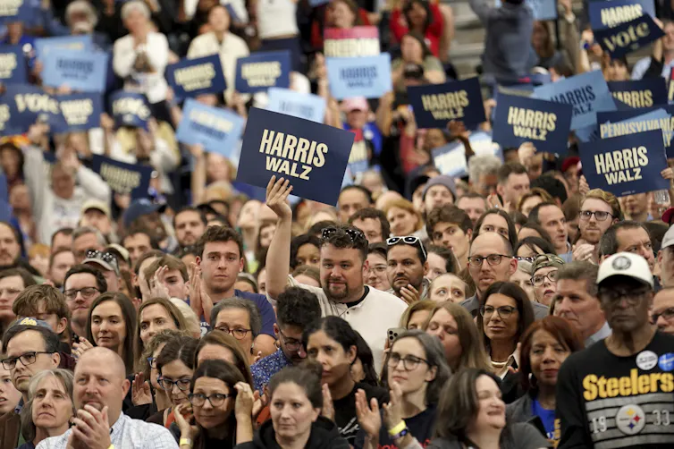 People hold Harris Walz signs. One man wears. Pittsburgh Steelers T-shirt.