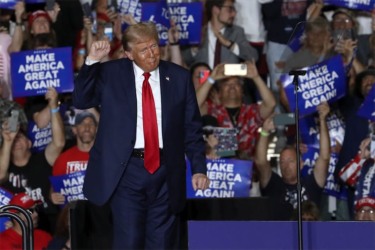 An overly tanned man with fluffy grey-blond hair in a baggy blue suit with a red tie gestures at a campaign rally, with his right arm raised.