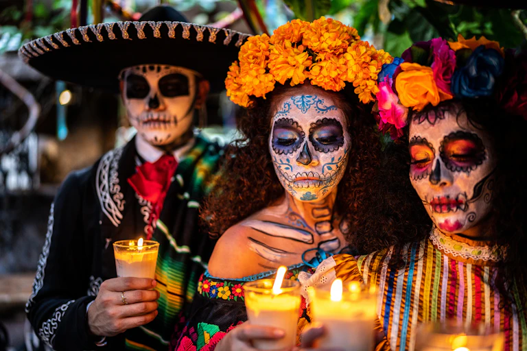 Three people with large hats and flowers around their heads hold candles, while wearing skull masks.