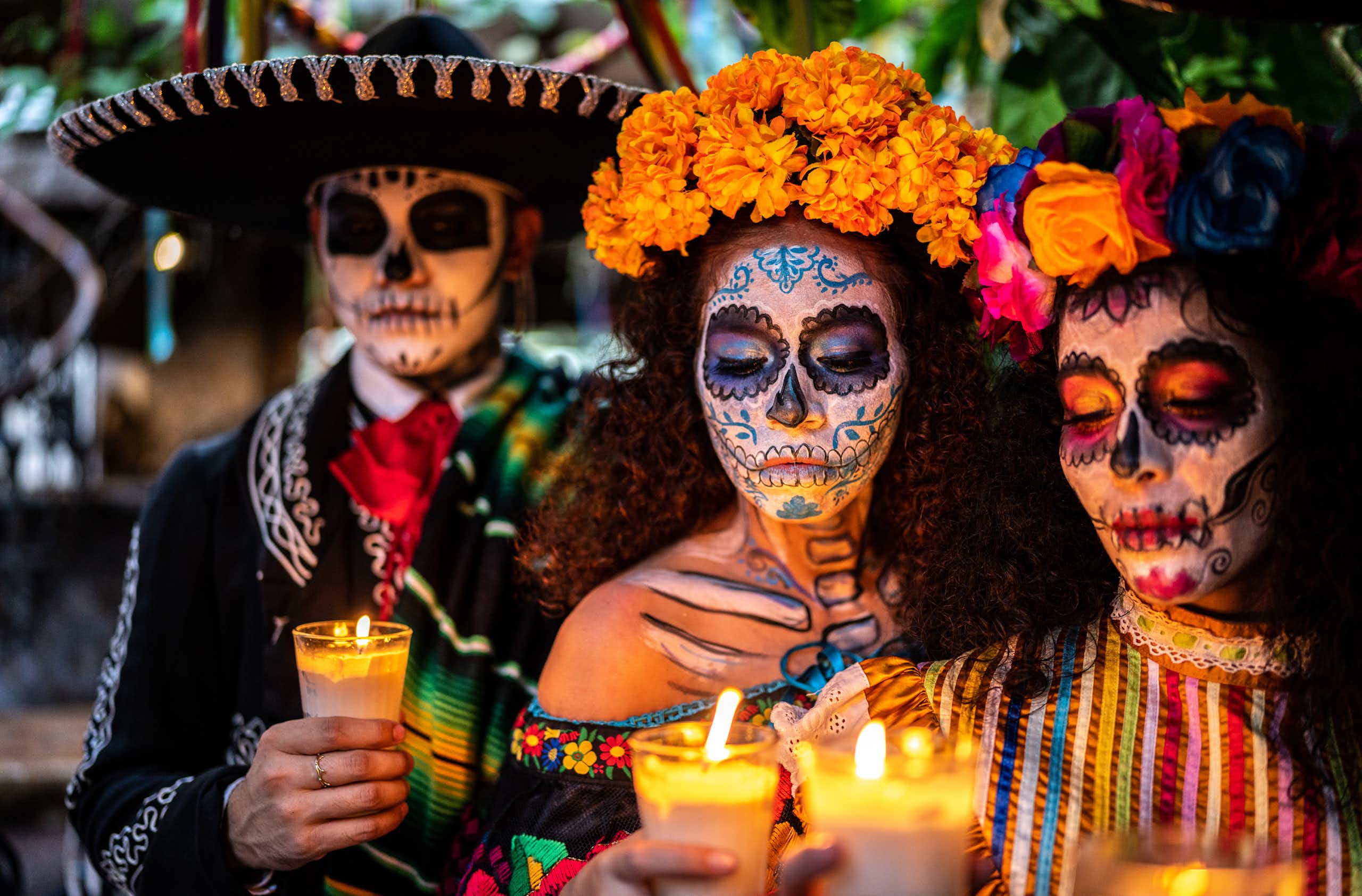 Three people with large hats and flowers around their heads hold candles, while wearing skull masks.