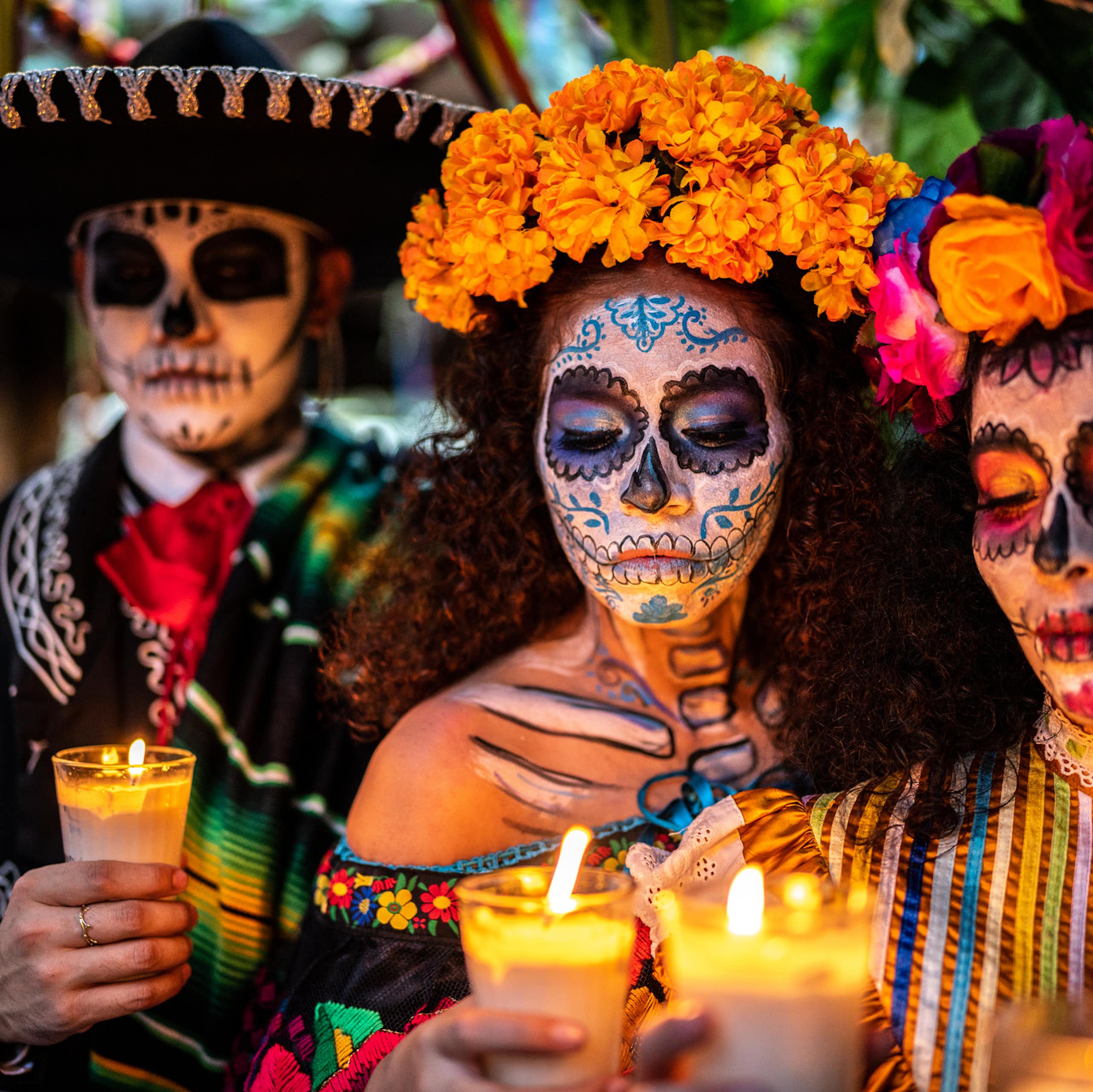 Three people with large hats and flowers around their heads hold candles, while wearing skull masks.