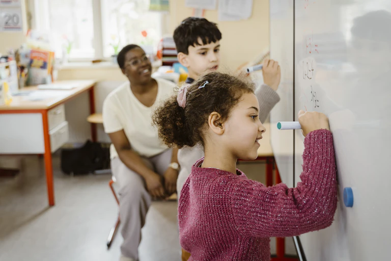 A girls and a boy use magic markers to do math problems on a whiteboard.
