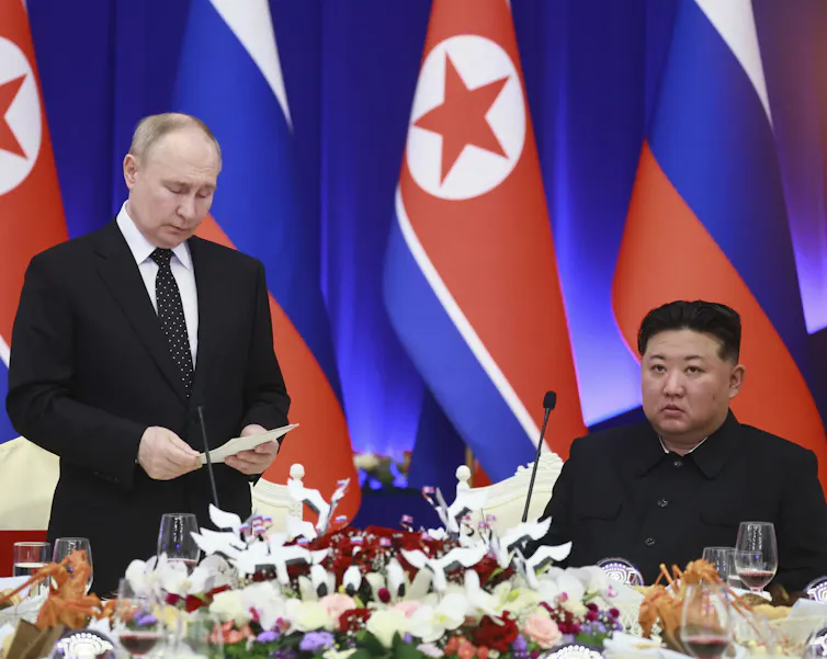 A balding white man speaks while looking at a document in his hand while an Asian man sitting down listens.