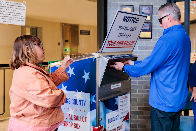 A voter dropping off a ballot in a large box with the American flag emblazoned on it.