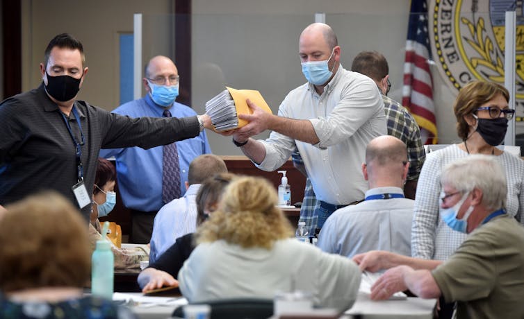 A group of people sitting and standing in a room that has an American flag on a pole, looking at pieces of paper.