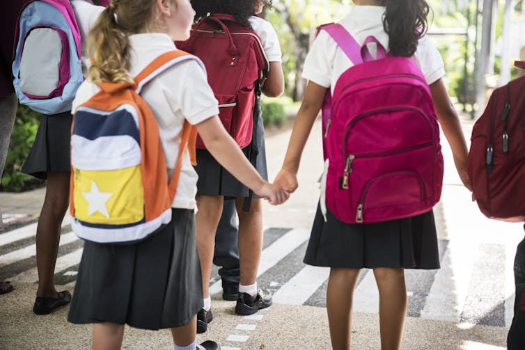 Young children in uniforms and backpacks cross a road.
