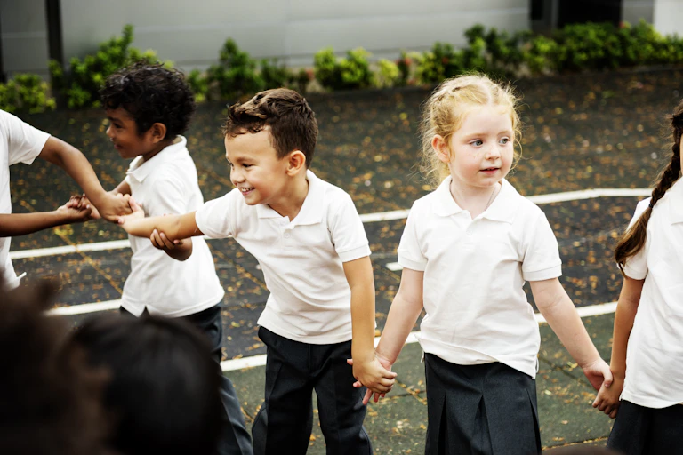 Young children in white shirts and dark pants hold hangs in a playground.