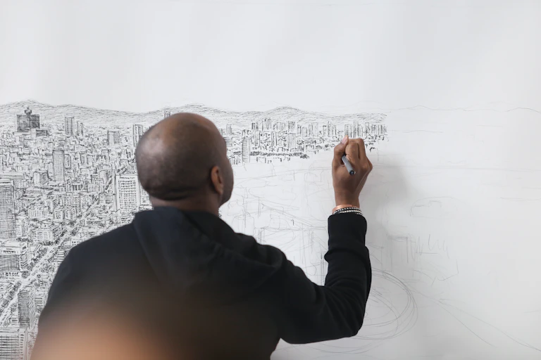 Stephen Wiltshire, de dos, dessine sur une grande toile tendue au mur le panorama de la ville de Mexico.