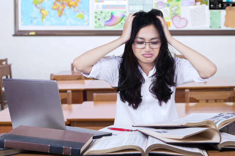 young woman head on her head, looking confused at desk of books and laptop