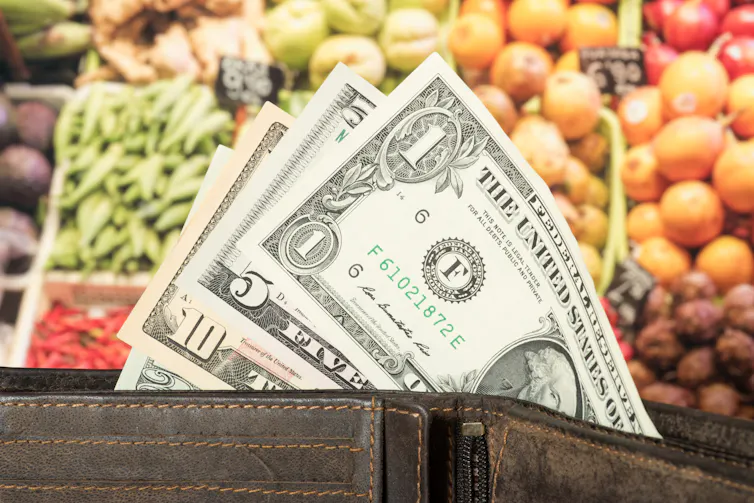 A wallet and US dollar bills in front of vegetables on a supermarket shelf.